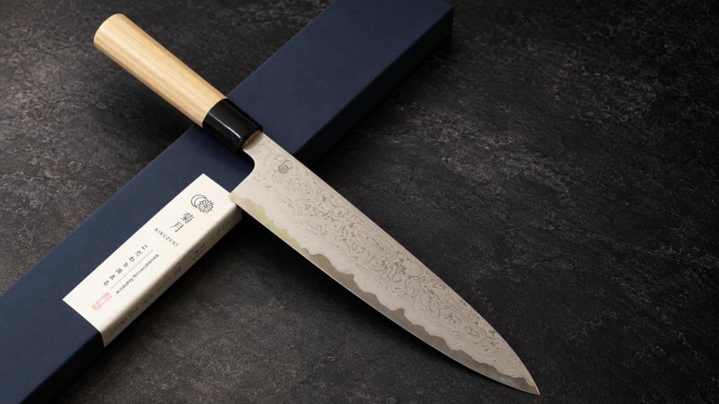 Sakai Kikumori Japanese gyuto knife with a Damascus-pattern blade and light wood handle displayed beside a dark presentation box.