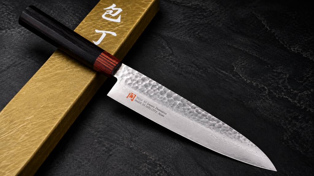 Iseya Japanese chef’s knife with a hammered Damascus blade and dark wood handle displayed beside a gold presentation box.