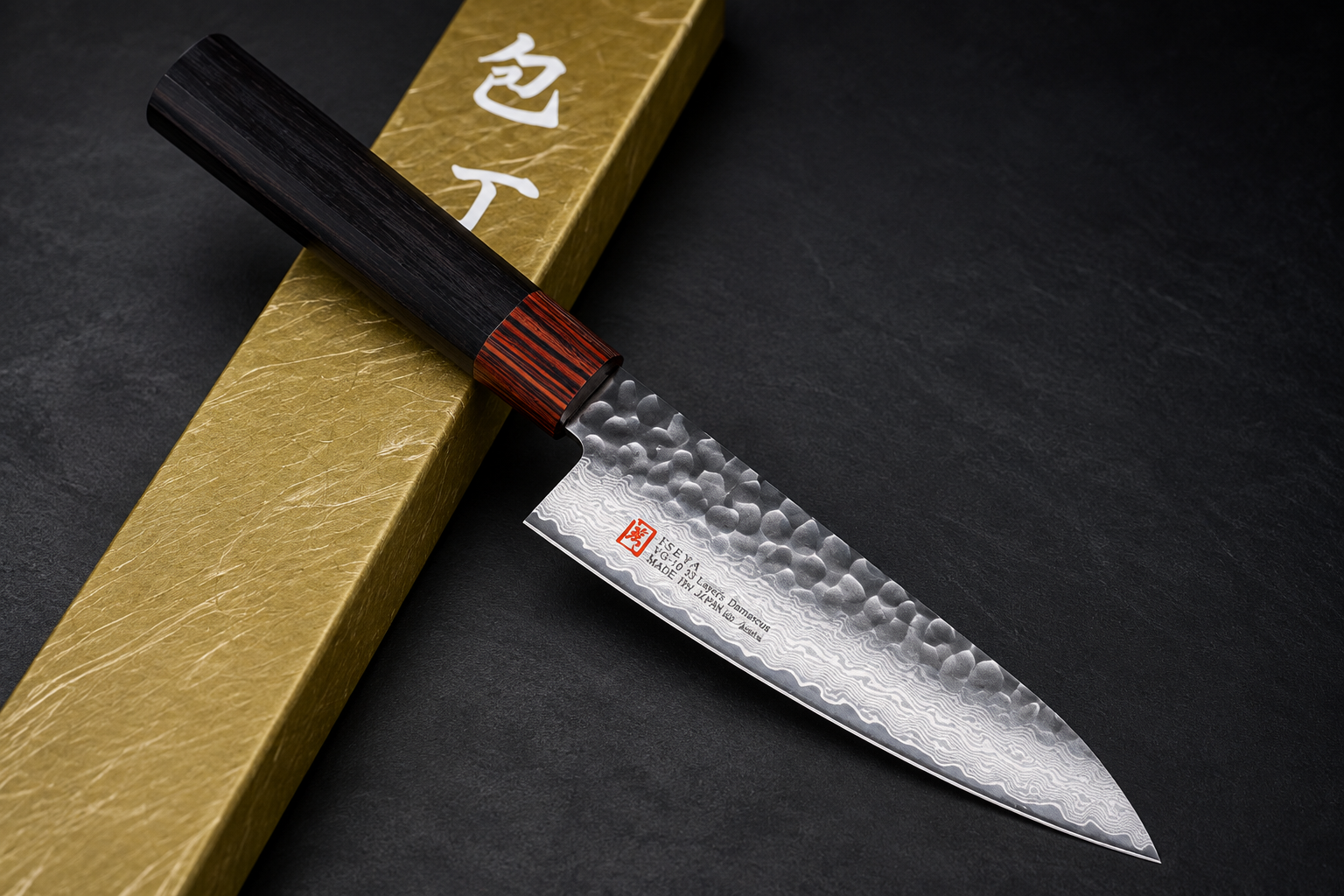 Iseya I-Series 33-layer VG-10 Damascus hammered Santoku 135mm with a dark wooden handle resting diagonally across a gold presentation box on a dark textured background.