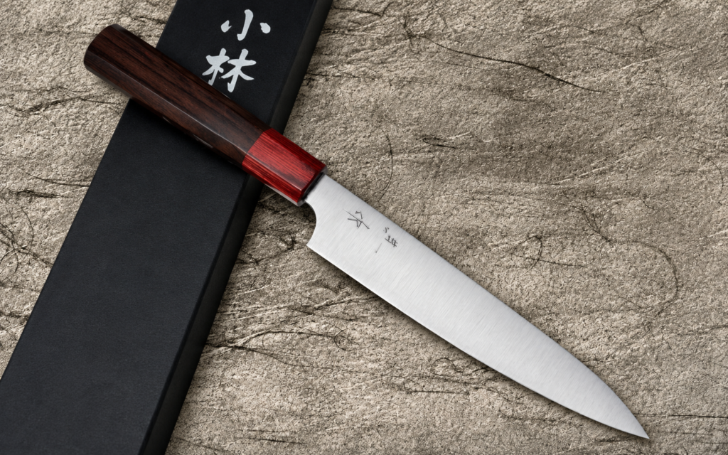 Kei Kobayashi R2 petty knife 150mm with a dark wood handle and red ferrule displayed beside a black presentation box on a textured surface.