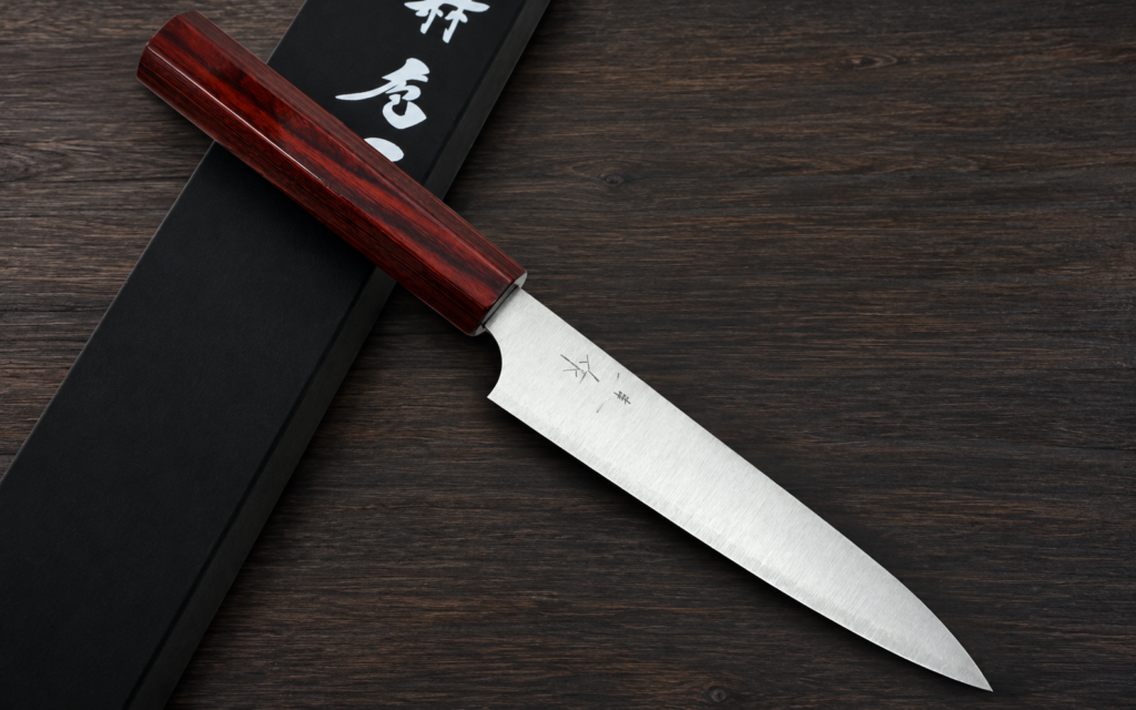 Japanese petty knife with a reddish-brown wooden handle and stainless steel blade resting diagonally over a black presentation box on a dark wooden surface.
