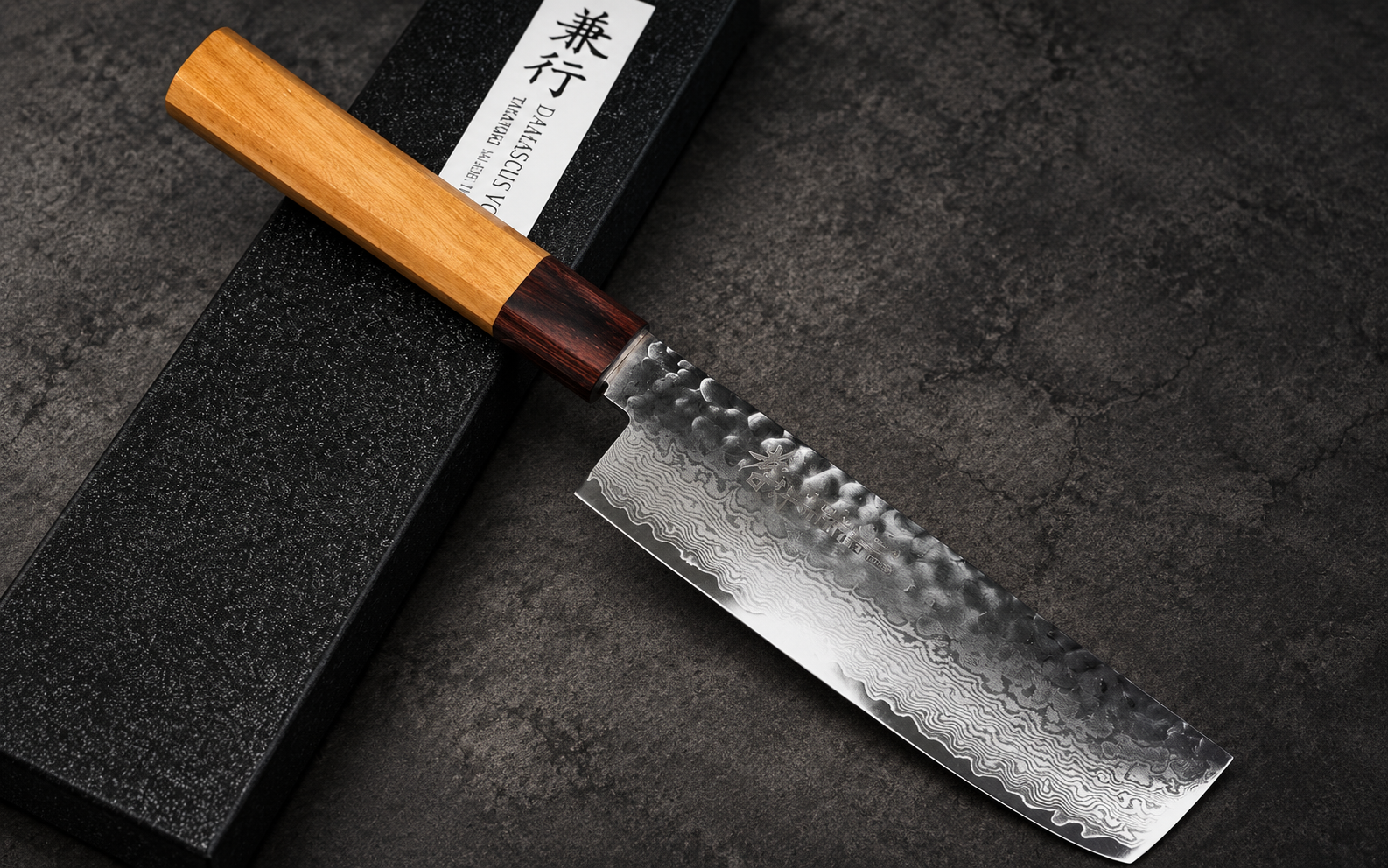 Sakai Takayuki 33-layer VG10 Damascus hammered Nakiri 160mm with a light wooden handle resting diagonally across a black presentation box on a dark textured background.