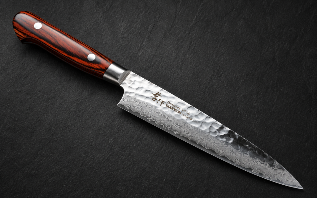 Japanese Damascus petty knife with a patterned blade and dark wooden handle on a dark stone background