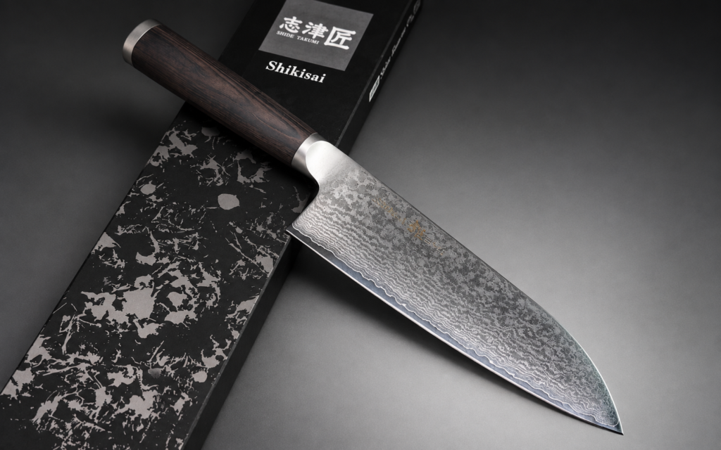 Shikisai MIYAKO 33-layer Damascus Santoku knife with a dark wooden handle resting diagonally across a black patterned presentation box on a dark gray background.