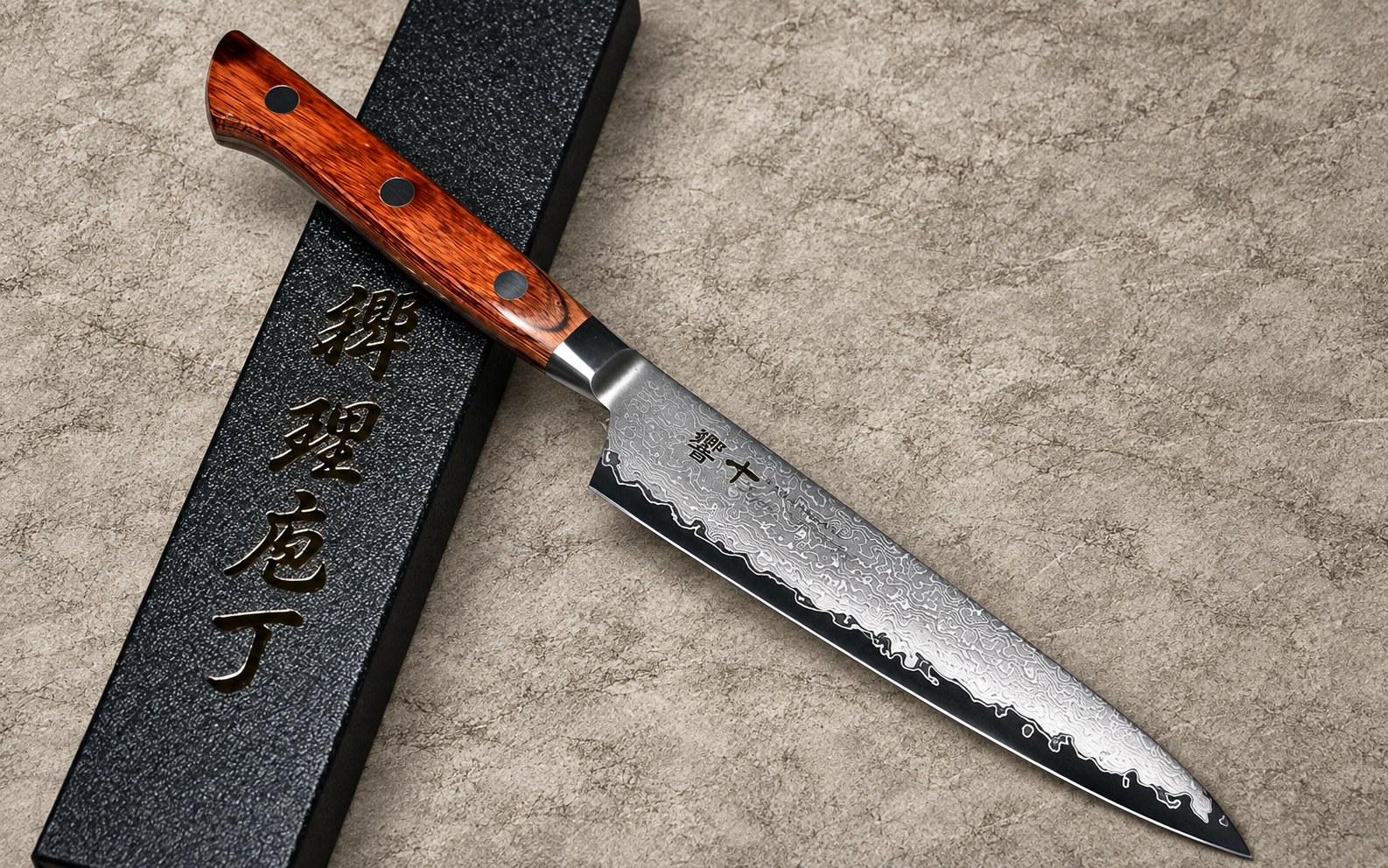 Tamahagane Kyoto 63-layer Damascus petty knife with a reddish-brown wooden handle resting diagonally beside a black presentation box on a textured beige background.