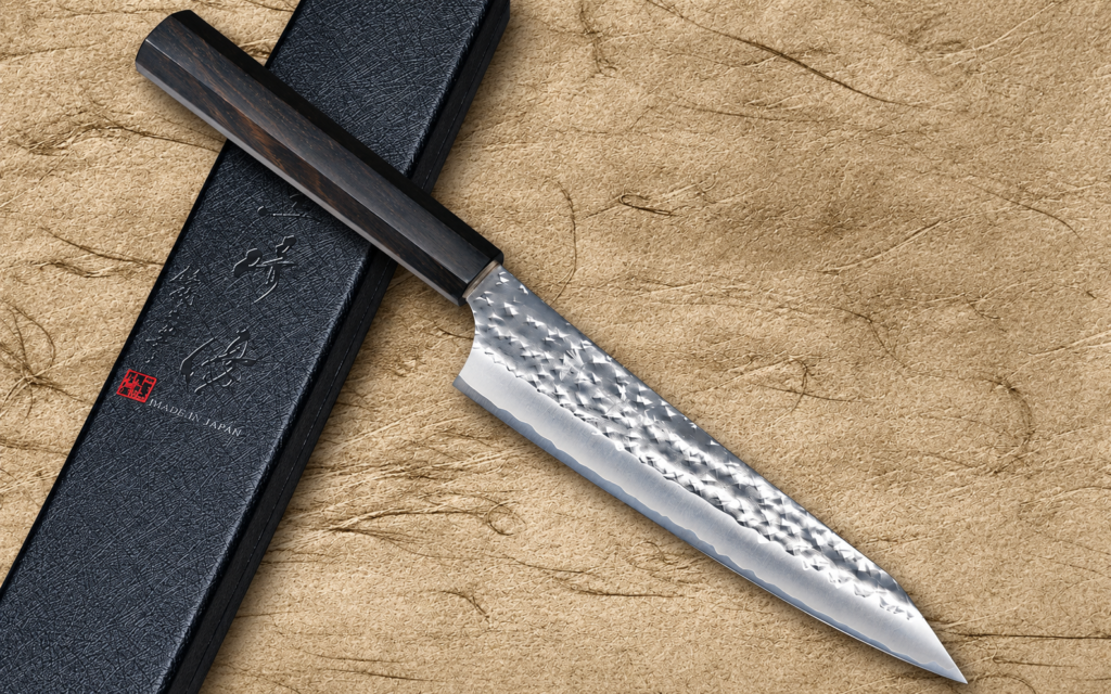 Yu Kurosaki Aogami Super Petty 150mm with hammered carbon steel blade and dark ebony handle beside a black Japanese knife box on a textured beige background.
