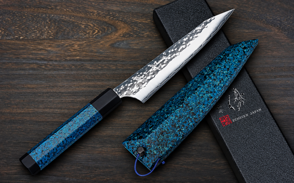 Yu Kurosaki R2/SG2 Petty 150mm with blue indigo handle and matching saya beside a black Echizen Japan box on a dark wooden surface
